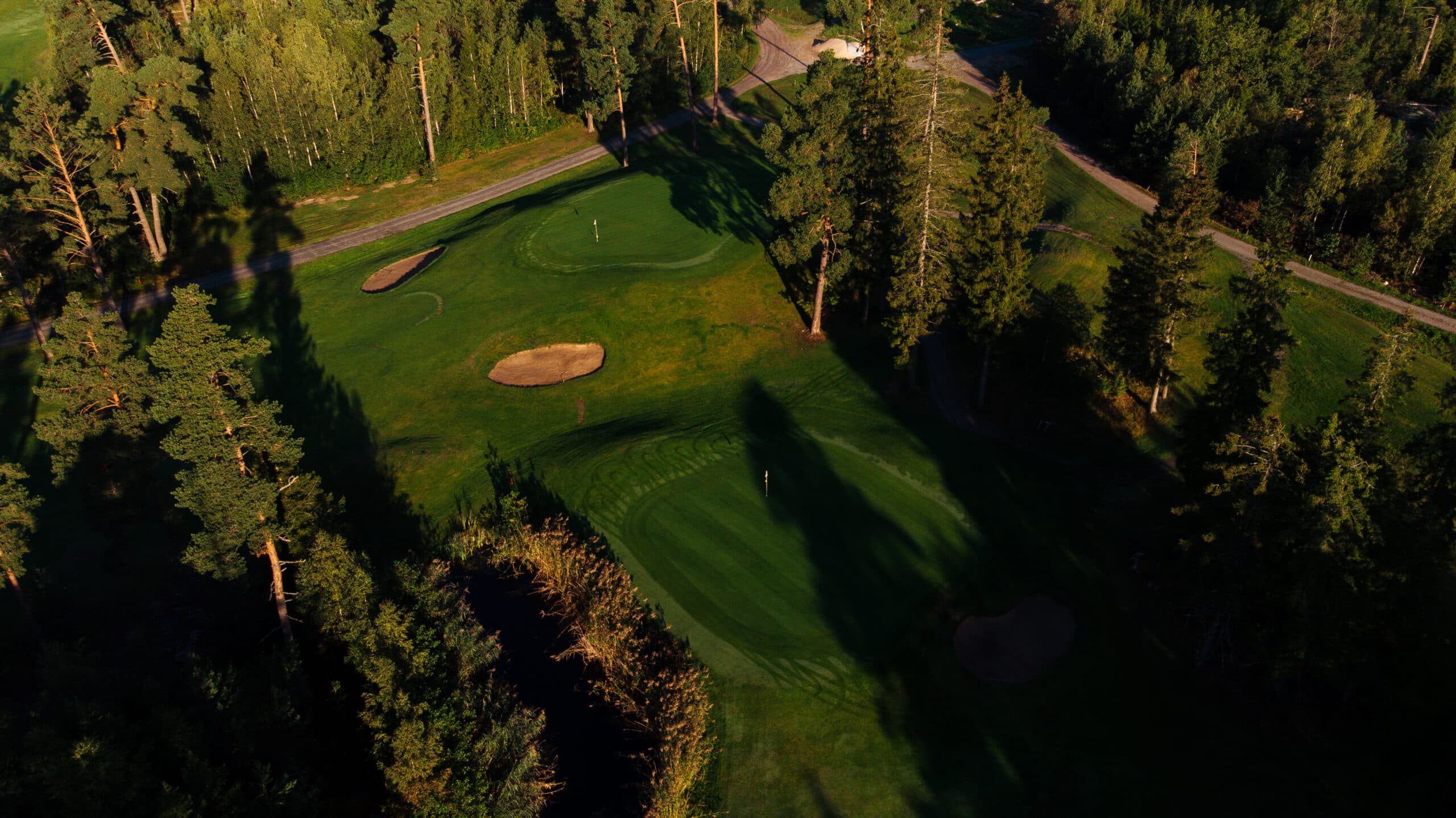 Flygvy över Ljusterö Golfs greener och bunkrar, omgivna av skog i kvällsljus