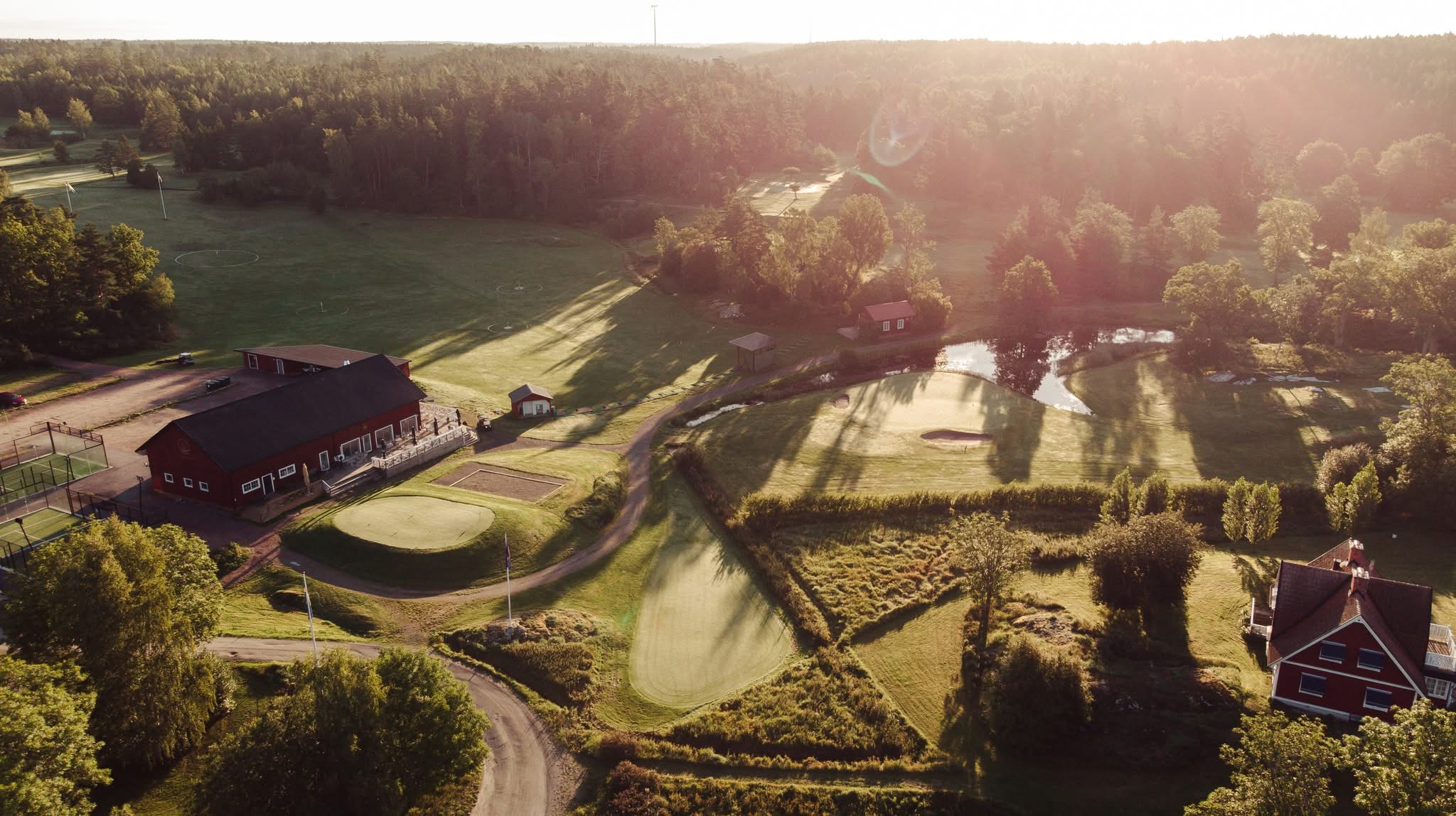 Flygbild i motljus över Ljusterö Golf, med drivingrangens övningsfält till vänster om klubbhuset